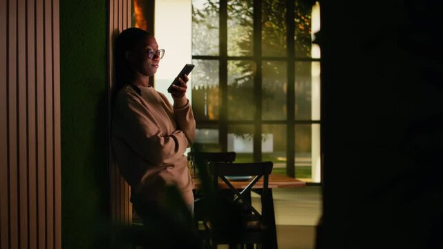 Young African American woman with phone sitting in the hallway in cozy ambiance, calm moment browsing mobile apps for web browsing. Warm lifestyle and casual screen time habits.