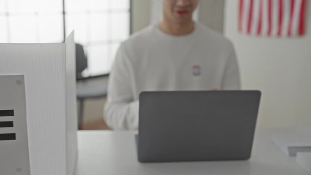 Young hispanic man wearing glasses with laptop and i voted sticker, hands gesturing above keyboard inside a voting booth building; civic pride.