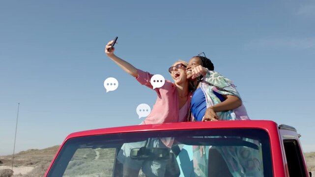 Extending phone, pink woman posing selfie; blue friend leaning in; viewing screen by dunes, red car