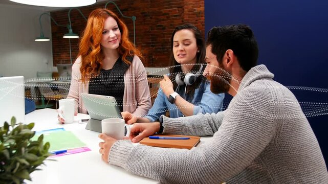 Discussing team sitting at meeting table, redhead in pink cardigan holding mug, man pointing tablet
