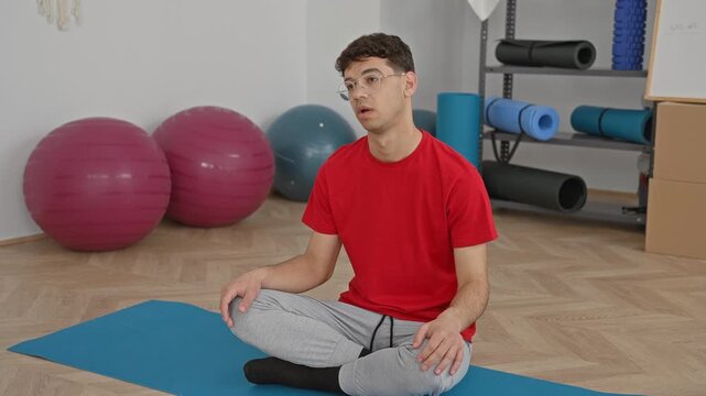 Man seated on yoga mat in studio with exercise balls and hand on forehead gesture showing tiredness; fatigue recovery.