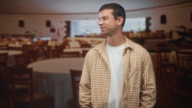 Man with glasses smiling, hands in pockets and wearing plaid overshirt inside a building dining hall; quiet contentment.