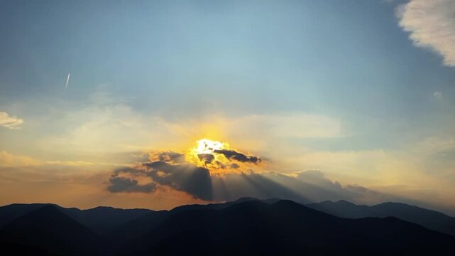 A cinematic 50-second long-take time lapse showing the complete process of a dramatic golden sunset with rays bursting behind clouds over layered mountain silhouettes.