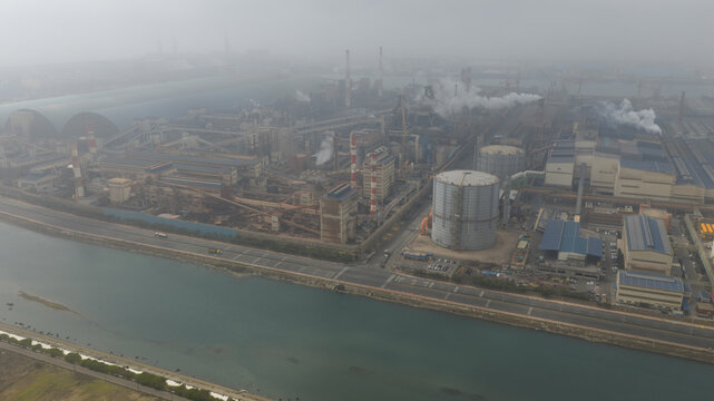Aerial view of the Taichung Power Plant with smokestacks emitting plumes of smoke and large storage tanks along a waterway under a hazy sky Lishui Village, Taichung City, Taiwan.