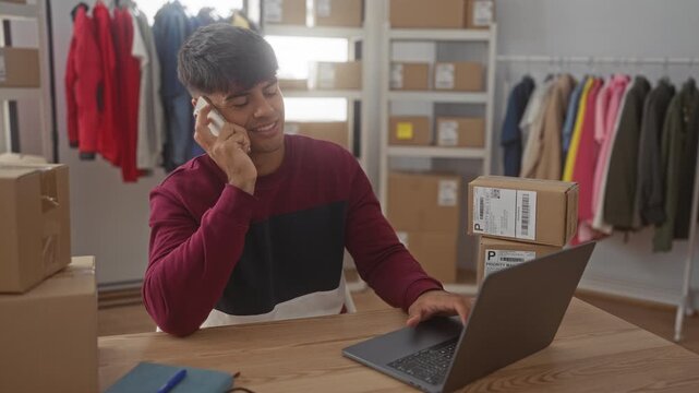 Man holds phone to ear and types on laptop amid stacked shipping boxes in studio workspace; focused small business hustle.