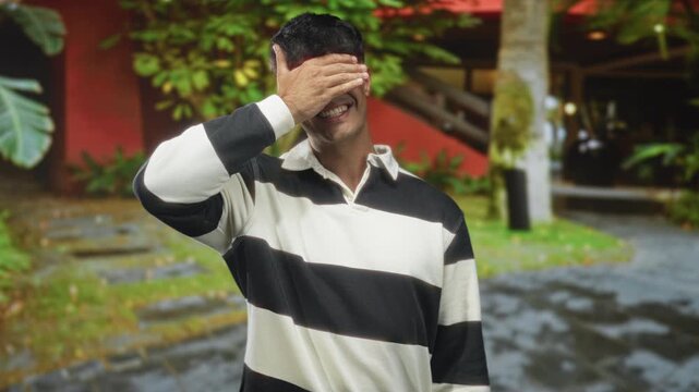 Man covering eyes with hand and smiling in front of building, wearing a striped shirt and casual collar; playful.