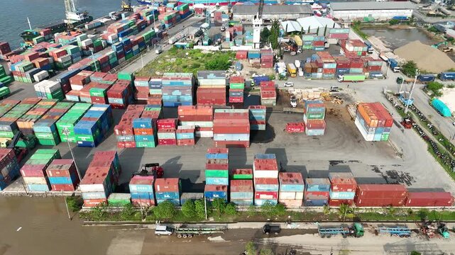 High angle aerial view of a reach stacker crane lifting a pink shipping container onto a heavy-duty truck trailer amidst rows of colorful stacked cargo containers at a busy industrial port.