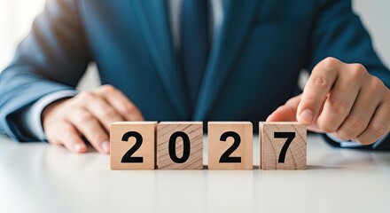 Obraz premium An executive in a dark blue suit rests their hands on a row of four wooden blocks displaying the future calendar year twenty twenty seven on a white desk.