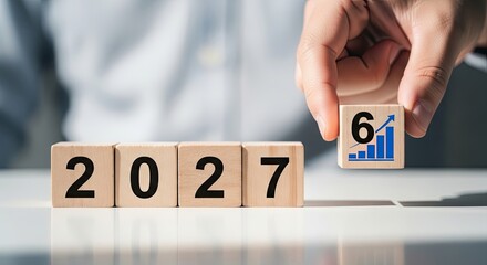Naklejka premium A person holds a wooden cube showing a blue upward growth arrow and the number six next to blocks displaying the year twenty twenty seven in an office.