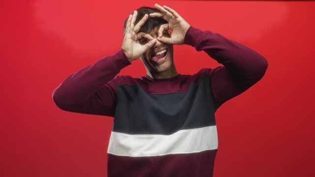 Young hispanic man with hands forming binocular gesture and tongue out in studio against red backdrop; playful fun.