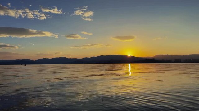 Cinematic 50 second time lapse of a golden sunrise reflecting on the calm water of Dianchi Lake with mountain silhouettes in Kunming China.