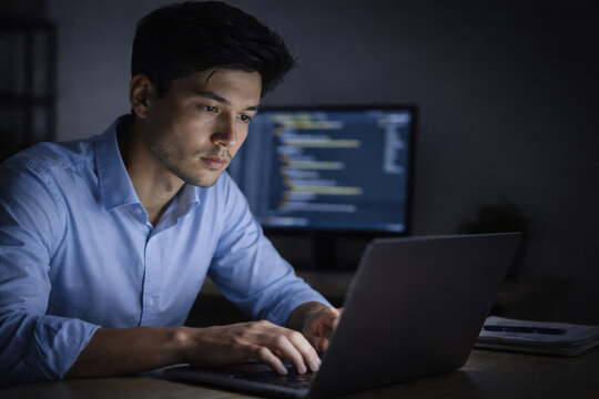 Focused male software developer programmer writing code on laptop in dark office environment with serious expression during late night work session