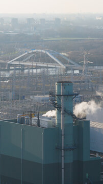 Aerial view of the Vattenfall Elektriciteitscentrale Diemen power plant with a smoking chimney, an arched bridge, and power lines under a hazy sky in Diemen, Noord-Holland, Netherlands.