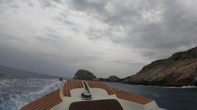 First-person POV view from a luxury motorboat with teak deck cruising through blue Mediterranean waters near dramatic rocky cliffs in Souda, Chania, Greece under dramatic cloudy skies.