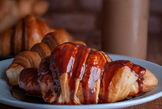 Croissants covered in melted dark chocolate on a white plate on a rustic wooden table with a glass of Fip Frappe Mocha latte cold coffee in the background. Close-up photograph, macro photography, hori