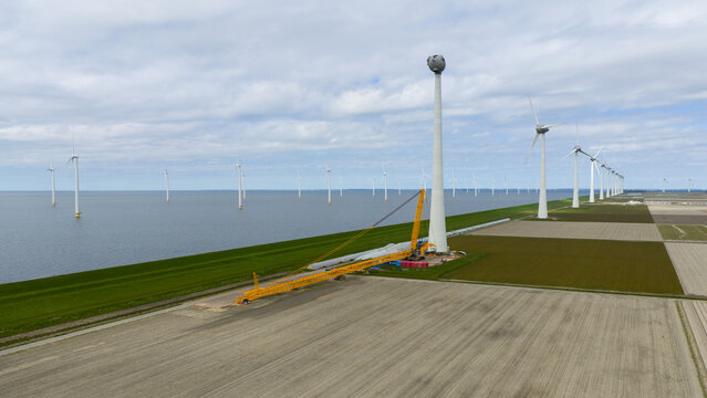 Aerial view of Westermeerdijk wind farm featuring a large yellow crane by a turbine tower, coastal fields, and offshore turbines under a cloudy sky in Espel, Flevoland, Netherlands.