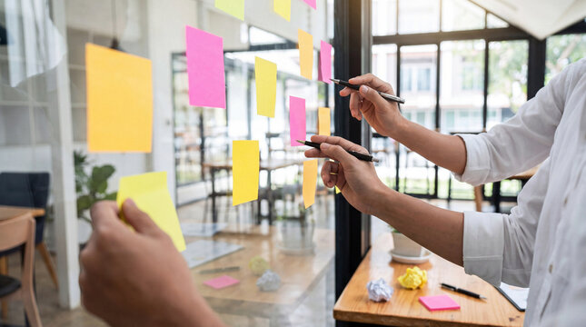 Creative professionals actively brainstorming and organizing ideas using colorful sticky notes on a clear glass board.
