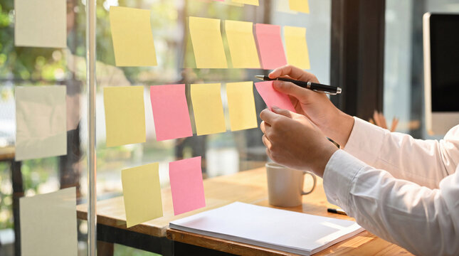 Person's hands organizing ideas on a glass board with colorful sticky notes in a modern office.