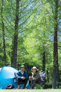 新緑のキャンプ場で椅子に座るカメラ目線笑顔のアウトドアやレジャーを楽しいミドルの夫婦全身の広角　上にコピースペースあり