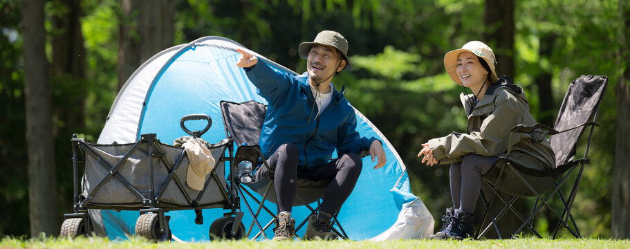 新緑のキャンプ場で椅子に座る笑顔のアウトドアやレジャーを楽しいミドルの夫婦　鳥や景色を眺める、指を差して見つめるバナー