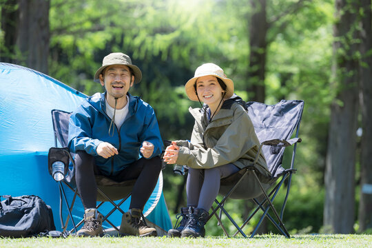 新緑のキャンプ場で椅子に座るカメラ目線笑顔のアウトドアやレジャーを楽しいミドルの夫婦