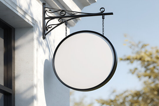 A blank, round sign hangs from a decorative bracket and chain on a building. The sky and tree foliage create a blurred background