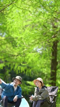 新緑のキャンプ場で椅子に座る笑顔のアウトドアやレジャーを楽しいミドルの夫婦　鳥や景色を眺める縦動画　指を差して見つめる