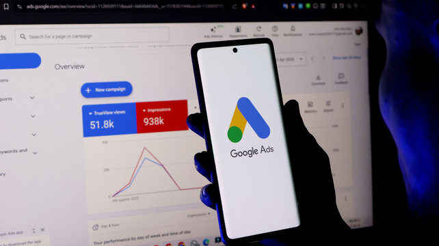 A person holding a smartphone with the google ads logo in front of a computer screen displaying an advertising dashboard