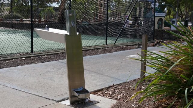 A stainless steel public drinking water fountain with a built-in dog water bowl at the base, on a concrete path beside a chainlink fenced tennis court in a suburban neighbourhood park in Australia