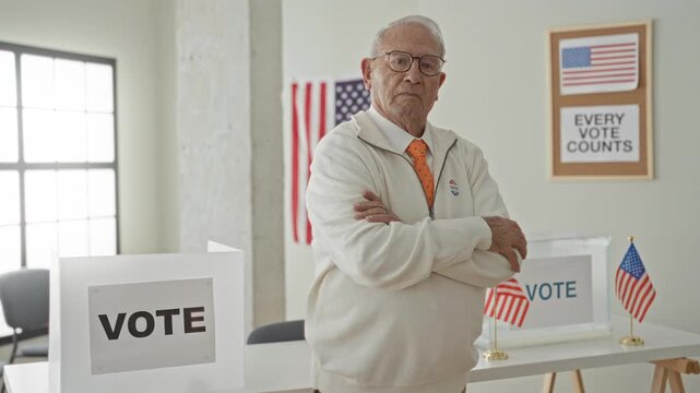 Senior man with arms crossed beside clear ballot box, vote sign and small american flags at a polling table in a public building; civic duty.
