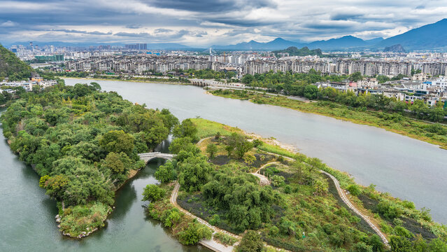 Urban landscape, view from a height. Lots of multi-storey buildings, green vegetation. In the foreground is the riverbed, an arched bridge over the water. Mountains on the horizon. Clouds in the sky.
