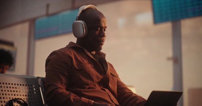 Professional African American Man Sits in Sun Drenched Airport Terminal, Preparing For Virtual Meeting. Male Wearing Headphones and to Join Business Conference. Concept Connected Corporate Lifestyle.