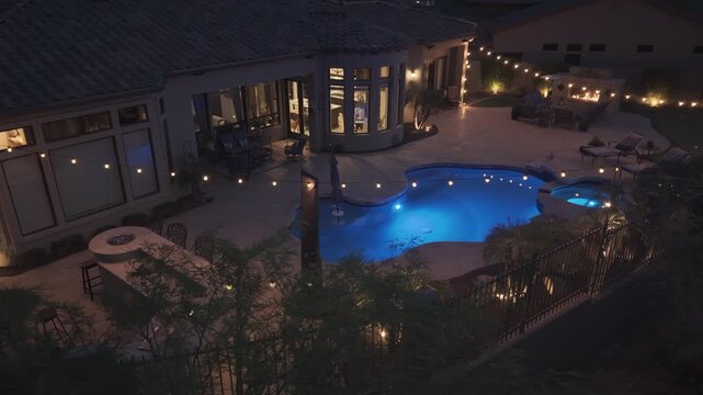 A 4k night aerial view of a desert landscaped home in Arizona featuring a travertine tiled pool deck with an outdoor fireplace and kitchen.