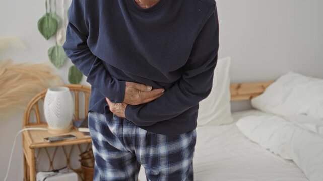 Man clutching abdomen with hands beside bed in building showing stomachache, wearing navy sweater and plaid pajama pants, bedside lamp and rattan nightstand visible; pain vulnerability.