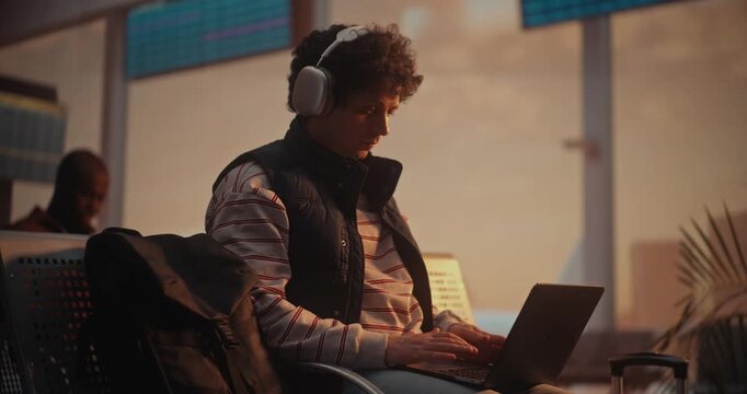 Young Man With Curly Hair Sits by Large Window in Airport Departure Lounge. Male Focused on Laptop Screen, Fingers Moving Rapidly Across Keyboard as Writes Code. Productivity of Modern Programmer.