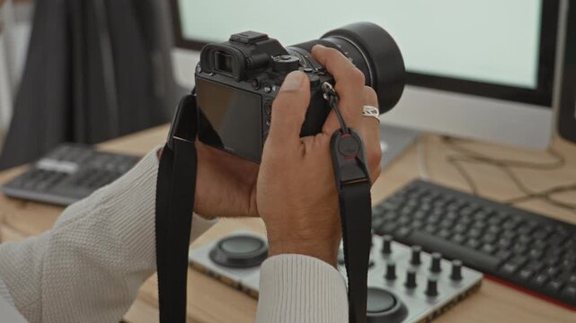 Man holding camera with fingers on shutter while adjusting lens in studio at a desk by a monitor and editing control pad, wearing white sweater; concentration craft skill.