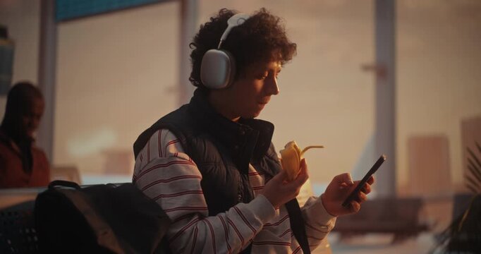 Sitting Comfortably In Departure Hall, Young Male Passenger Scrolls Through Social Media Feed and Eating Banana. With Headphones on, Man Immersed In Digital World, Oblivious Busy Airport Environment.