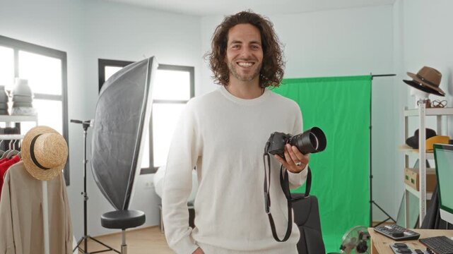 Young hispanic man holds camera in hand and smiles in a photo studio with green backdrop and softbox visible; confidence creativity.