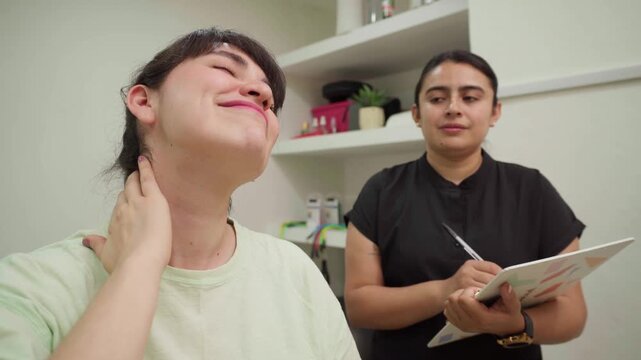 Relieved patient smiling and massaging her neck during a medical consultation with a physiotherapist taking notes in a modern physical therapy clinic office