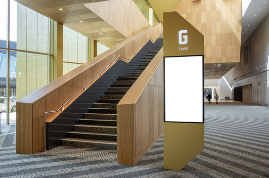 A blank vertical digital signage mockup on a sleek pylon, situated next to a contemporary wooden staircase in a spacious public foyer. Background texture for  advertising and wayfinding
