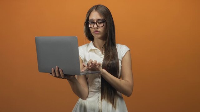 Woman holding laptop and typing with fingers in studio against an orange wall, wearing glasses and white sleeveless dress; focus.