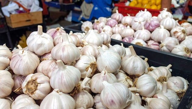 Close-up shot of multiple fresh garlic bulbs piled high within black containers, showcasing a market scene with other produce visible