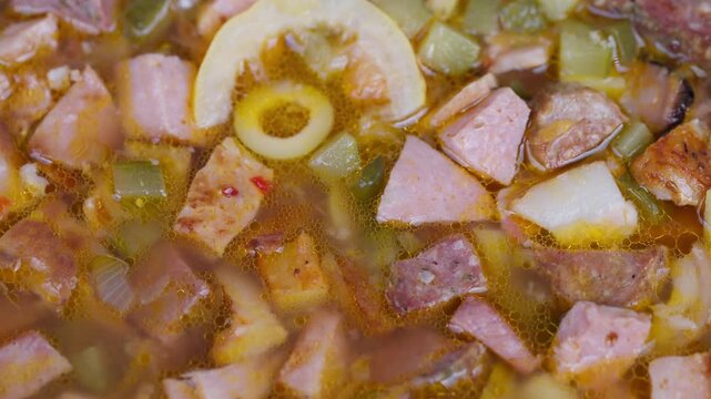 Closeup homemade soup with diced sausage and potato cubes, olive slice and pickle pieces in golden broth flecked with red pepper, glossy texture and rustic cozy mood