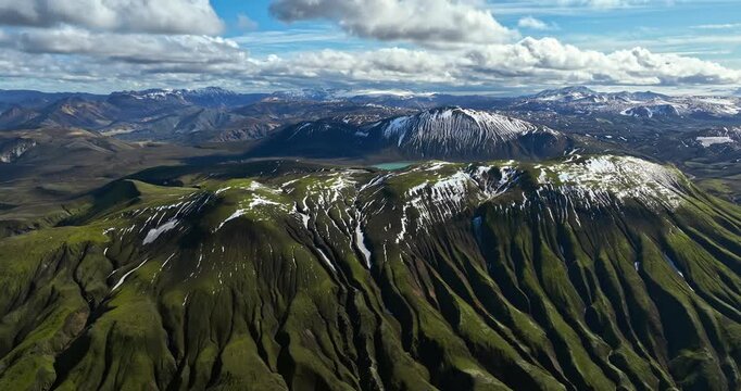 Aerial view of dramatic green volcanic ridges streaked with snow, revealing remote highlands, deep erosion lines, and vast mountain wilderness under textured clouds.