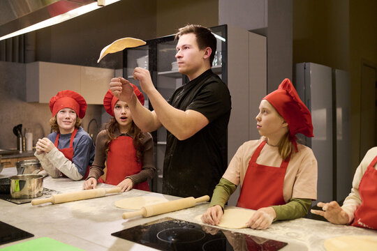 Young adult Caucasian man teaching children tossing pizza dough in cooking class, learning teamwork and food skills. Useful for culinary education, family activity marketing