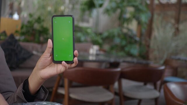 Woman holding up a smartphone with a green screen, showing tracking markers, sitting in a cafe.