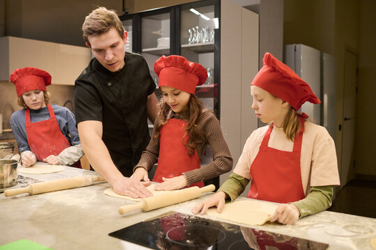 Caucasian male chef guiding kids rolling dough during cooking class in training kitchen, supporting teamwork and practical learning. Useful for culinary education, parenting, youth programs