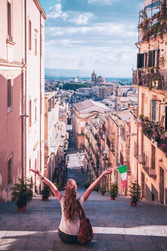Young woman sitting on Santa Maria del Monte stairs holding Italian flag, looking at Caltagirone cityscape. Travel lifestyle and freedom concept in Sicily, Italy. Famous ceramic stairs view.