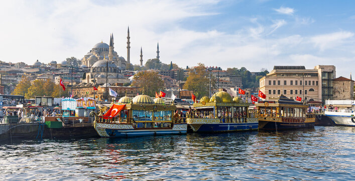 Traditional Ottoman style fish boats in Eminonu with Suleymaniye Mosque in background, Istanbul, Turkey. Golden Horn waterfront with Turkish flags and floating restaurants at sunny day.