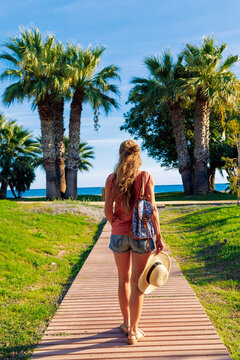 Young woman walking on wooden path or paseo toward to the beach between palm trees. Summer travel lifestyle and vacation concept with sea view, straw hat and backpack. Tropical coastal landscape at su